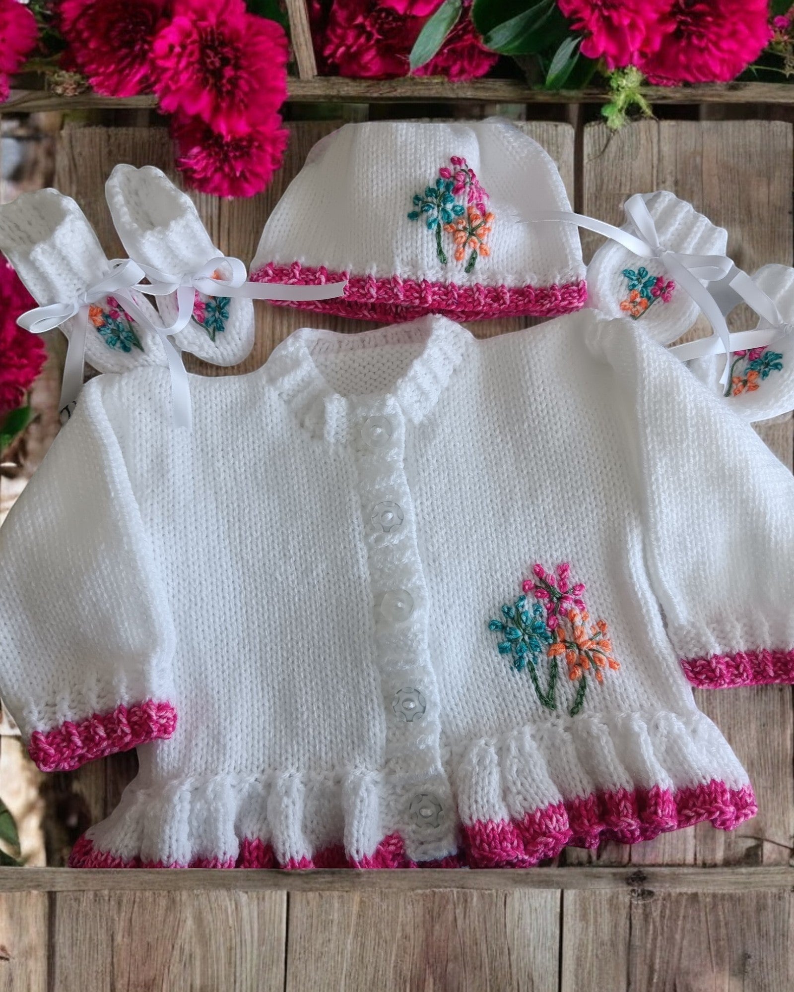 gemknits4kids baby girl wearing a white hand-knitted cardigan with floral embroidery, matching hat, and soft booties.
