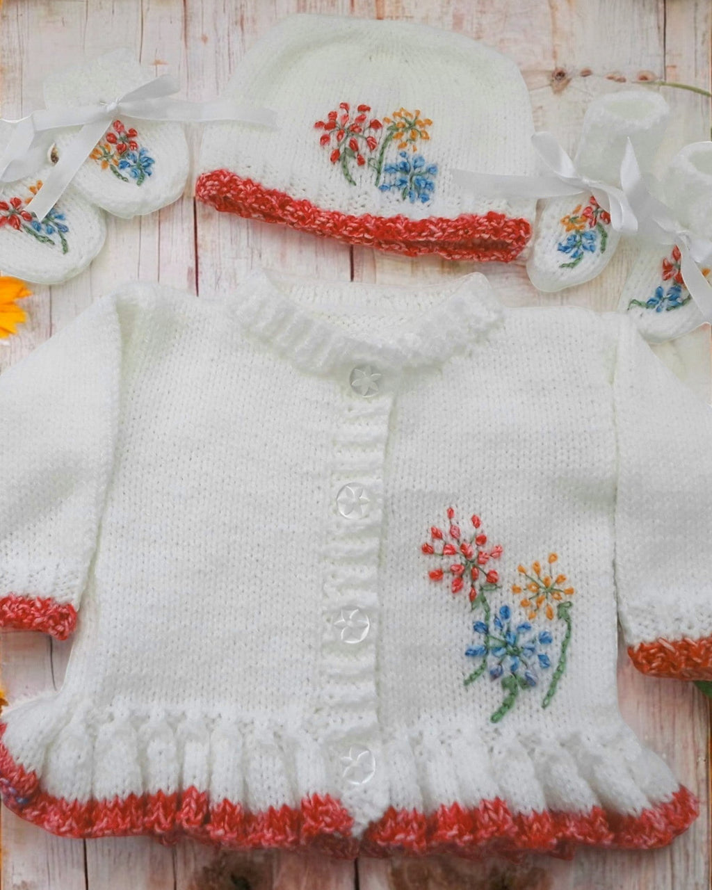 gemknits4kids baby girl wearing a white hand-knitted cardigan with floral embroidery, matching hat, and soft booties.