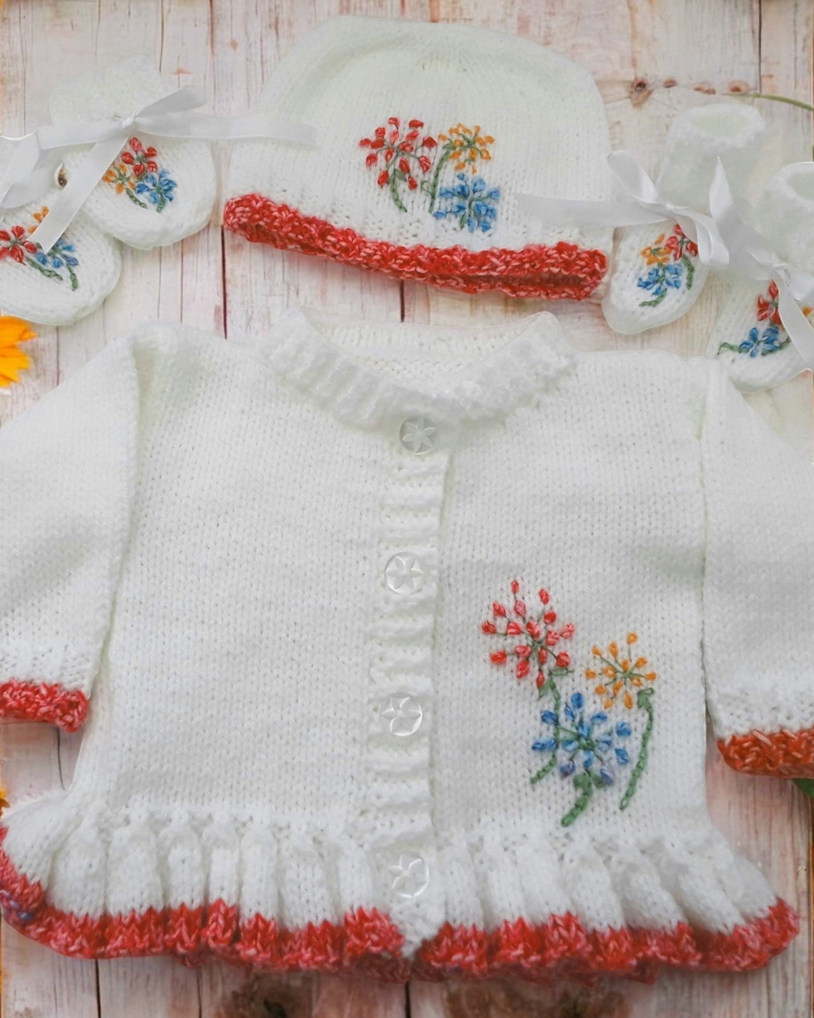 gemknits4kids baby girl wearing a white hand-knitted cardigan with floral embroidery, matching hat, and soft booties.