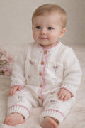 Baby girl wearing a gemknits4kids cream and pink knitted romper with hand-embroidered flowers and front button fastening.