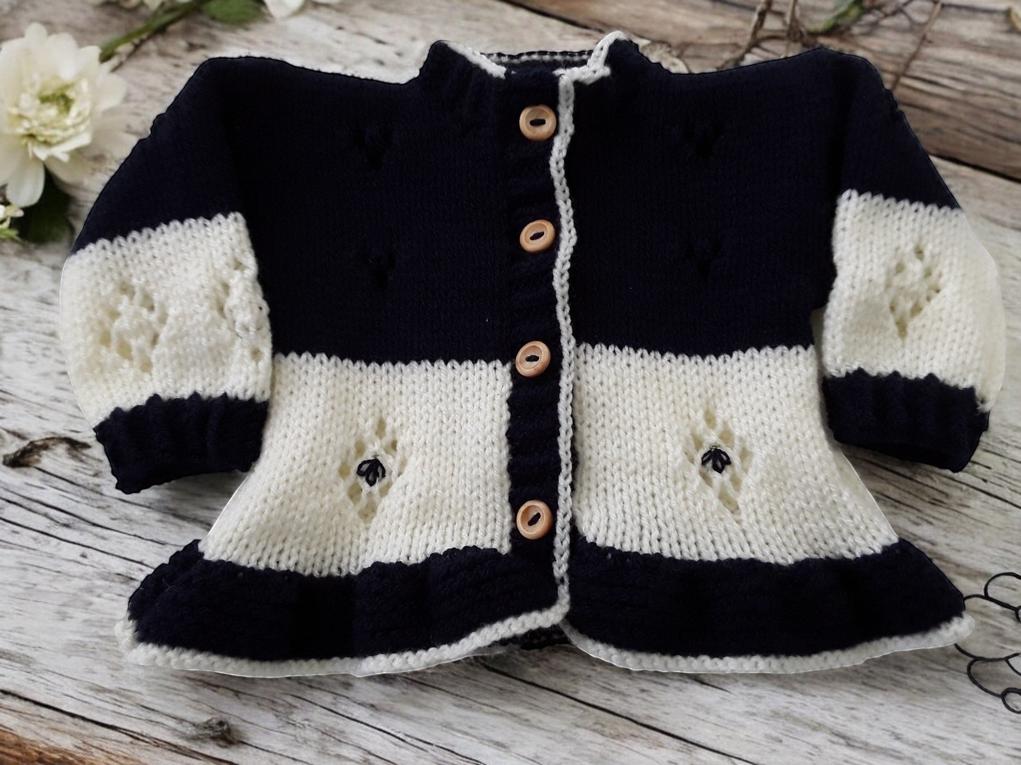 gemknits4kids baby girl wearing a two-tone navy and cream hand-knitted frilly cardigan, bonnet, and mittens set.