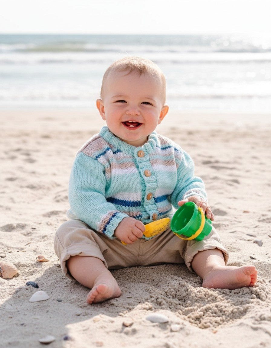 gemknits4kids baby boy wearing a pastel striped hand-knitted cardigan with wooden buttons, sitting on a sandy beach.