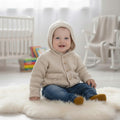 gemknits4kids baby wearing a soft beige knitted hooded jacket with buttons and pockets, sitting on a white rug.