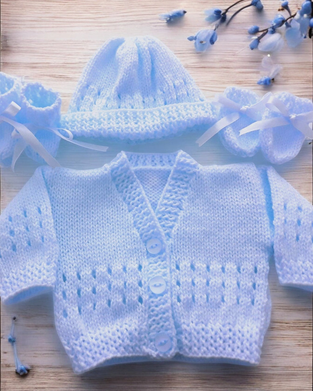 gemknits4kids baby wearing a soft blue hand-knitted cardigan, hat, and booties set sitting on a white rug in a bedroom.