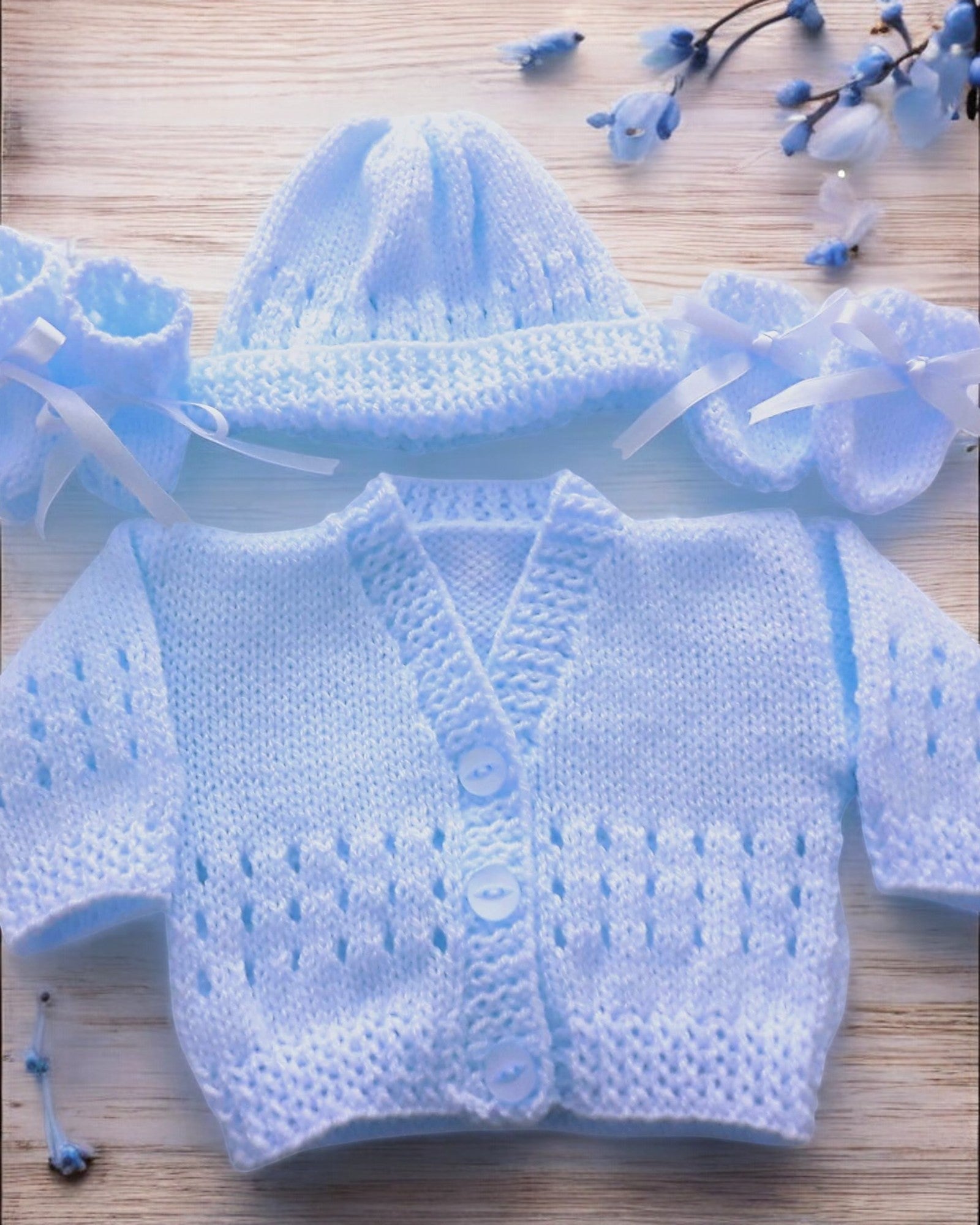 gemknits4kids baby wearing a soft blue hand-knitted cardigan, hat, and booties set sitting on a white rug in a bedroom.