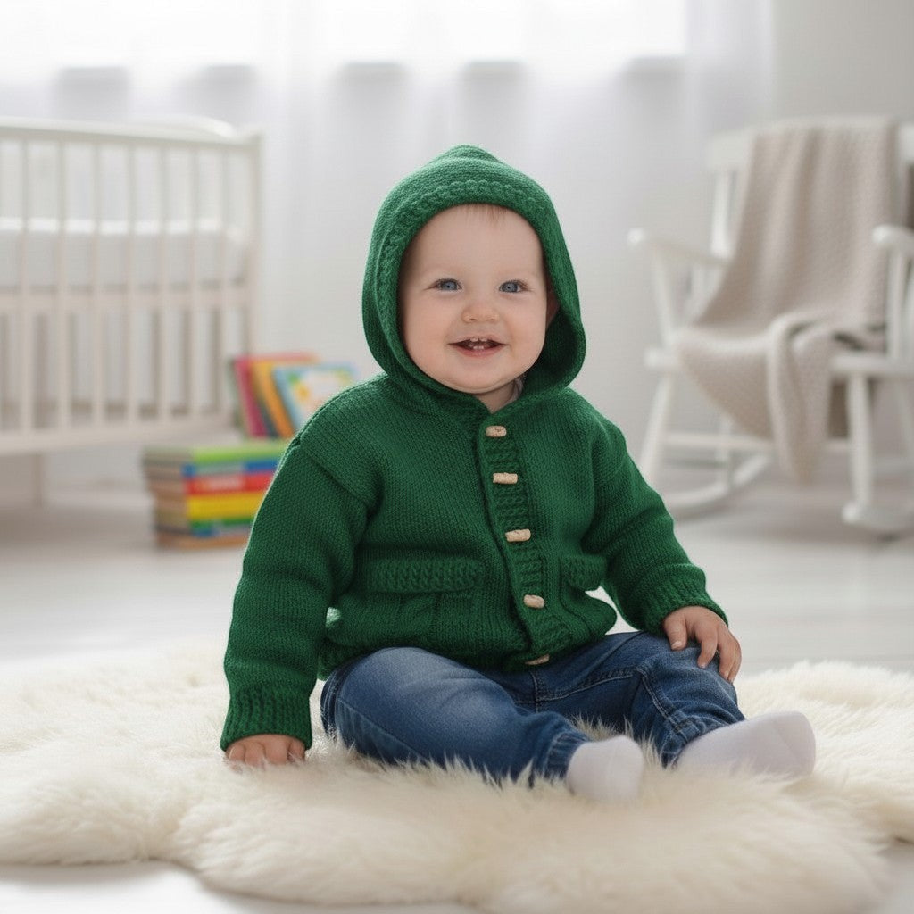 gemknits4kids baby wearing a soft green knitted hooded jacket with buttons and pockets, sitting on a white rug.