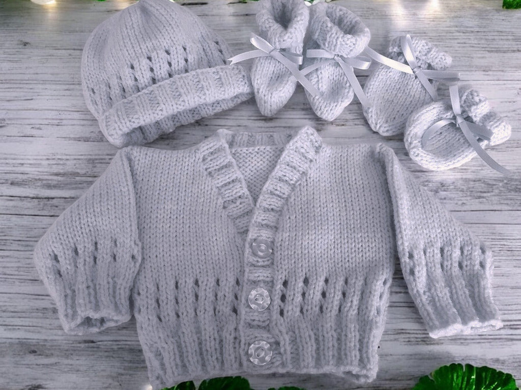 gemknits4kids baby wearing a soft silver hand-knitted cardigan, hat, and booties set sitting on a white rug in a bedroom.