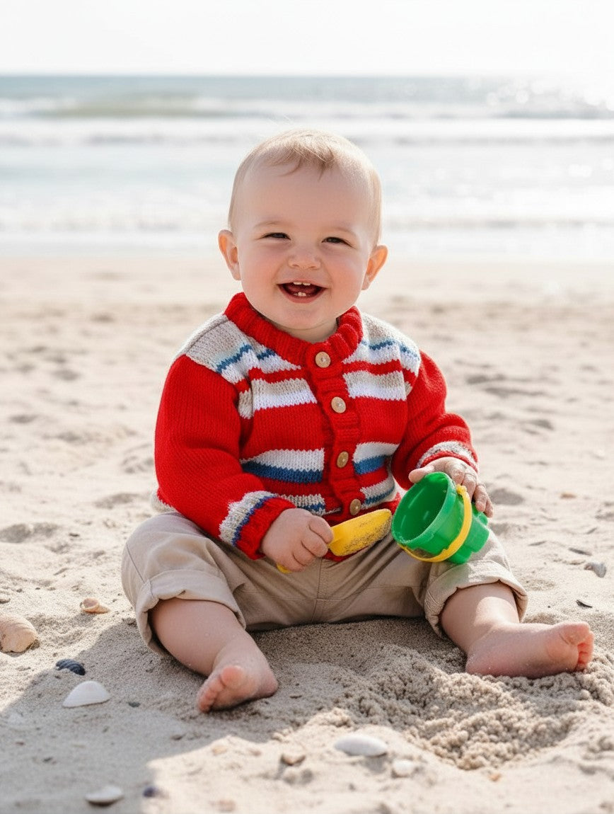 gemknits4kids baby boy wearing a red, white, and blue striped knit cardigan with wooden buttons on a sandy beach.