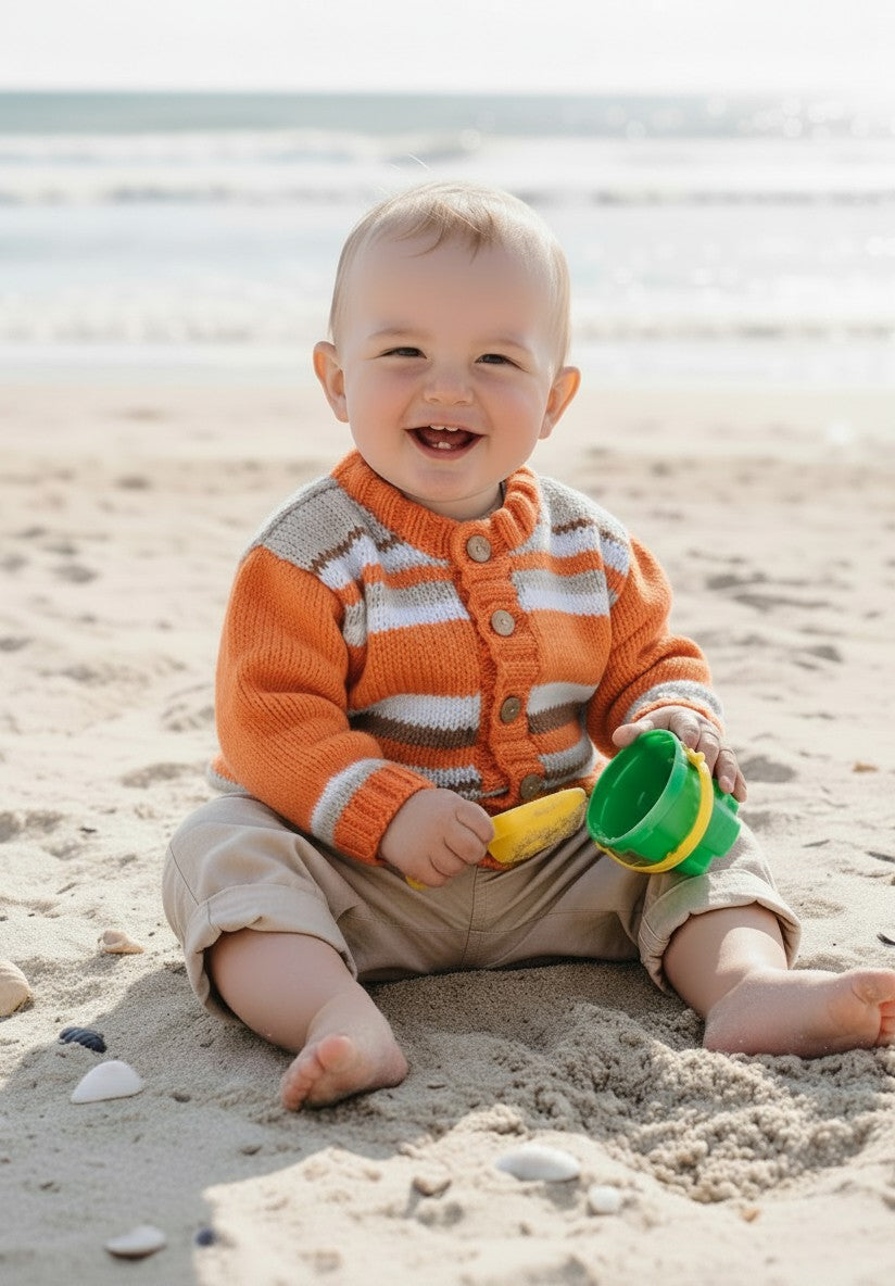 gemknits4kids baby boy wearing an orange and white striped hand-knitted acrylic cardigan with wooden buttons on a beach