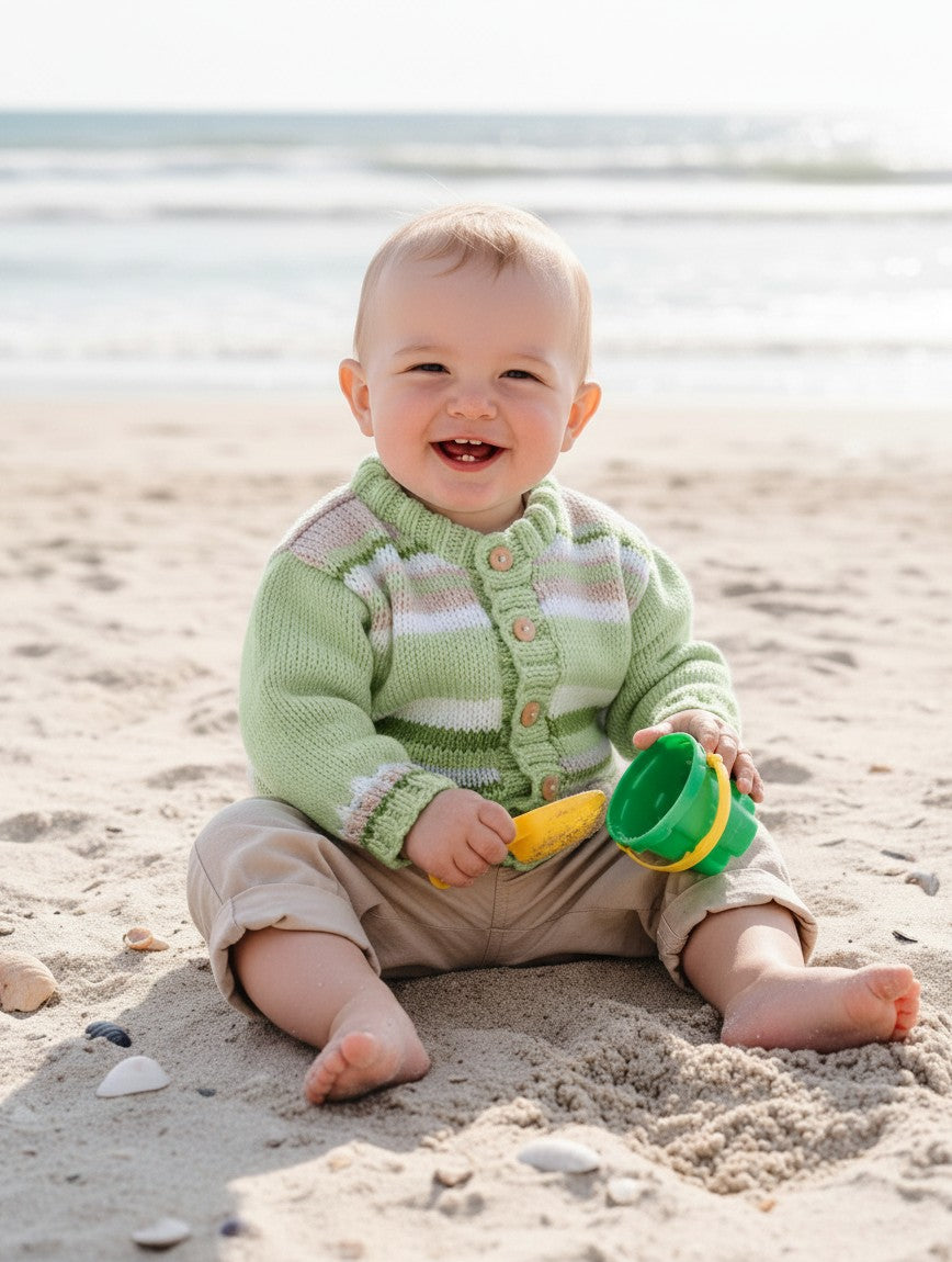 gemknits4kids baby boy wearing a hand-knitted green and white striped cardigan with wooden buttons on a sandy beach