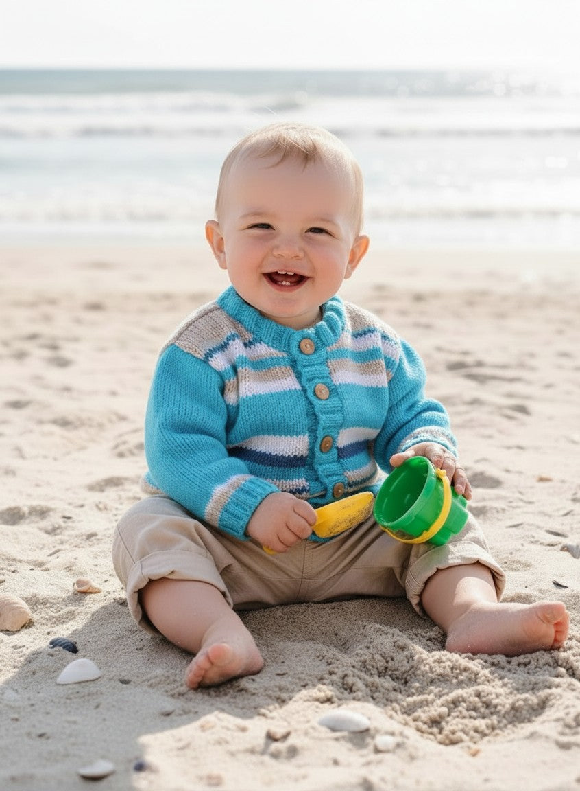 gemknits4kids baby boy wearing a blue and white striped hand-knitted acrylic cardigan with wooden buttons on a beach