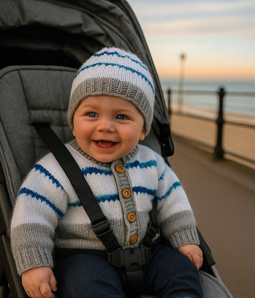 gemknits4kids baby boy wearing a hand-knitted Grey, blue & white striped cardigan, matching hat, and booties in a pram by the seaside